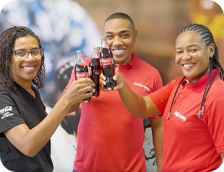 Tres colaboradores sonrientes realizando un brindis con botellas de vidrio de Coca-Cola, representando la cercanía y generación de valor social en las comunidades.